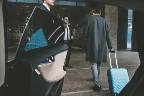 Businessmen in formal attire exiting a car, one of them carrying a blue wheeled bag, en route to the airport for an airport transfer.