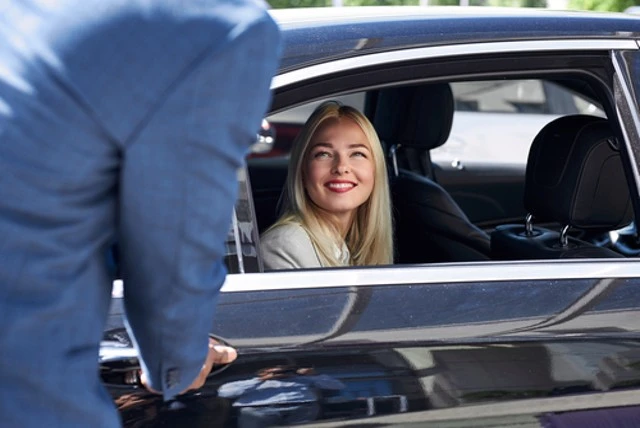 A blonde Caucasian woman, providing customer service, smiles at a black male as she sits in the driver's seat of a private car for a long distance ride.