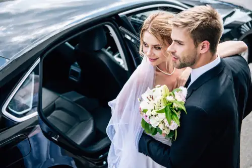 Elegant bride and groom by black car. Groom in suit hugs blonde bride with bouquet. Lavish ride chauffeur service in Houston.