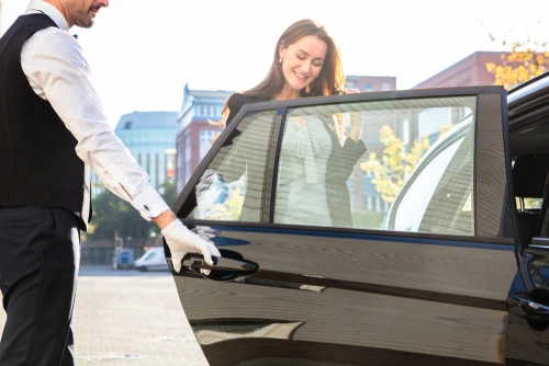 A professional chauffeur in uniform opens the door for an elegantly dressed, smiling young woman entering the back seat of a black luxury sedan, symbolizing Houston to Galveston luxury transportation.