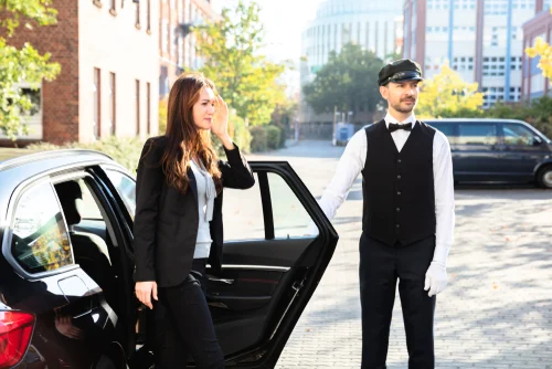 A professional chauffeur opens the rear passenger door for a stylish young businesswoman stepping out of a luxury black car—symbolizing Lavish Ride’s premium Galveston cruise shuttle service from Houston.