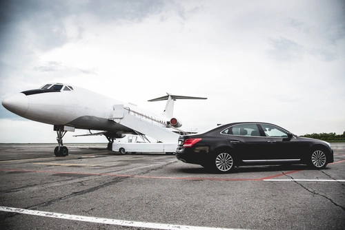 A black luxury car waits on the airport tarmac beside a private jet, ready for a businessman to disembark. This represents private jet ground transportation in Houston.