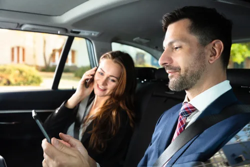 A businessman and businesswoman sit in the back seat of a black luxury car; she talks on her smartphone while he shows her something on his device, symbolizing the Houston Hobby Airport (HOU) private chauffeur service.