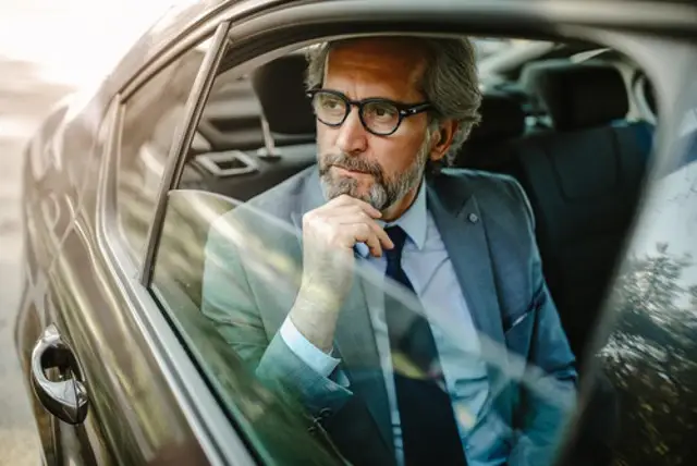 A man in a formal suit looks out of the open window of a black luxury car while using the hourly chauffeur service.