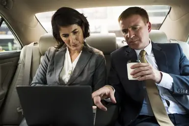 During their corporate executive transportation, a CEO and an employee share the back seat of a luxury black sedan; she works on her laptop while he drinks coffee and gives her notes.