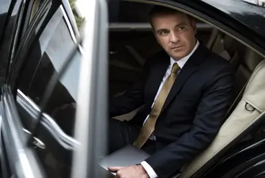 A businessman in formal attire holding a MacBook prepares to exit a luxury black car after an airport chauffeur service.