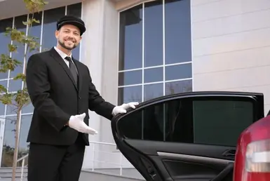 A professional luxury car driver warmly opens the back door of a black car for a customer with a smile.