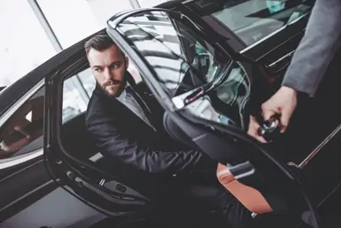 An elegant, uniformed chauffeur holds open the back door of a black car, assisting a businessman as he exits after using corporate chauffeur services.