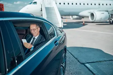 A businessman smiling while using his mobile phone in the back seat of a black car departing the airport runway, with a plane parked on the tarmac behind him, after using a private chauffeur service.