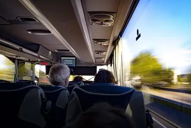 An interior view from the rear of a luxury passenger bus shows several passengers and part of the exterior, representing social group transportation services.