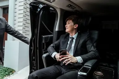 A young man dressed in a suit and tie sits comfortably inside a luxury black van while the driver holds the door handle, closing it as part of a professional point-to-point car service.
