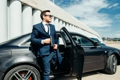 A young businessman in a suit and sunglasses, holding a laptop, exits a black car from a Corporate RoadShows.