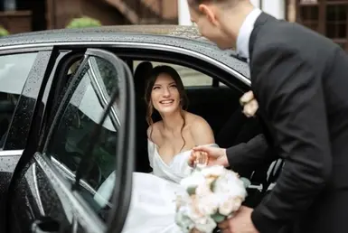 A suit-clad groom holds a bouquet and helps his bride in a white dress exit a luxury sedan at their wedding venue, using wedding limo services.