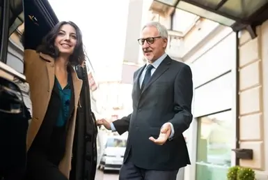 A professional driver helps a woman exit a black luxury van after reaching her destination using airport limo service, holding the door open with one hand and waving goodbye with the other.