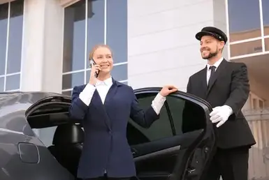 A professional driver opens the door of a black sedan limousine for a smiling businesswoman exiting the vehicle, representing black car ground transportation services.