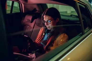Businesswoman in glasses using smartphone in back seat of chauffeured car during nighttime FBO airport transfer, showcasing executive private jet ground transportation.