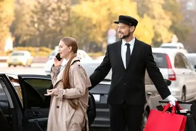 A professional driver opens the door of a black sedan limo for a young lady who is about to get in. She is talking on the phone while he carries her bags to begin the Birthday Party Limo Service.