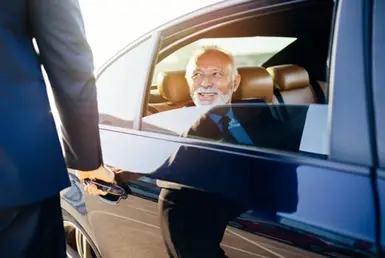 A professional chauffeur assists an elderly man in a suit as he exits a car, likely on his way to a medical appointment. Symbolizing our Expert Elderly Transportation Services.