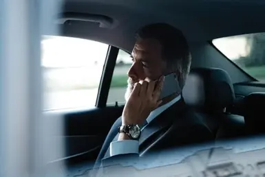 An elderly man in a suit and tie sits comfortably in the back seat of a car, talking on a cell phone, highlighting the vehicle's comfortable interior during long-distance elderly transportation.