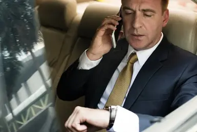 A businessman sits in the back seat of a luxury black car, speaking on his phone while a professional chauffeur drives by, symbolizing the Houston roadshow transportation service for corporate travelers.