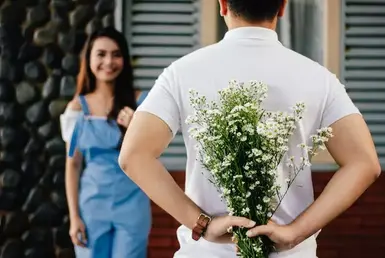 A man carries a bouquet behind his back in front of a happy woman standing near a marble wall.