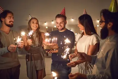 A joyful group of young adults wearing colorful party hats celebrates a birthday at home, holding glowing sparklers and smiling as they gather around a cake with lit candles. Earlier, they arrived using a birthday limo services in Houston.