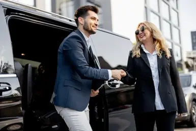 As the businessman exits the limo, a female chauffeur greets him. This represents a luxury taxi service for business people is with personal drivers. It is provided by Houston VIP Transportation service for Insurance Corporate.