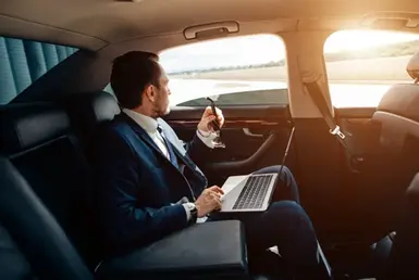 A businessman sits in the back seat of a luxury black sedan, using his Mac computer and looking out through the open window, enjoying the scenery. This symbolizes the private car transportation services to the NRG Stadium.