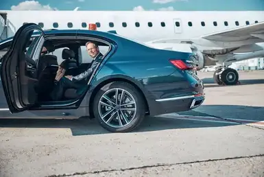 A smiling, elegantly dressed man with glasses in the back seat of a chauffeured car, departing from the airport tarmac—illustrating private car service to NRG Stadium.