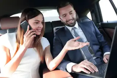 A business man and woman are seated in the back seat of a luxury black car; he is showing her something on his laptop while she is talking on her cell phone, representing event transportation services in Conroe.