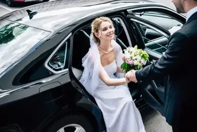 A suit-clad groom holds the hand of his elegantly attractive bride, helping her out of a wedding getaway car while she holds a bouquet of roses in his other hand.