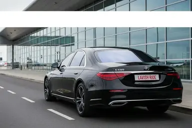 A Mercedes-Benz S-Class sedan, specifically an S 500, with the license plate "LAVISH RIDE," drives past a large building, symbolizing Mesa Airport Car Service.