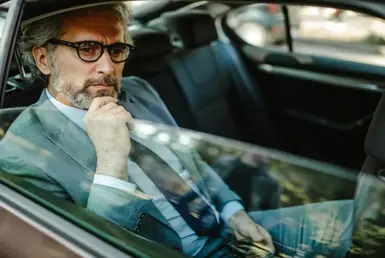 A businessman in a suit and tie observing city streets from the backseat of a car during a long distance ride.