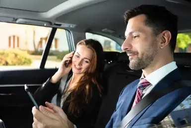 An elegant businessman and woman sit in a luxurious black car; she's talking on her phone while he's checking some of his work on his, symbolizing the luxury car service in Houston.