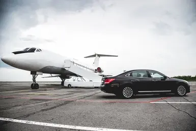 A black luxury sedan stands on the tarmac with a parked plane visible behind it, symbolizing bush airport to galveston.