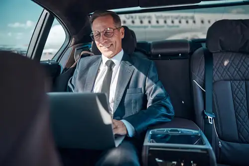A businessman is inside a luxury black car, leaving the airport while talking on his phone and smiling. A plane parked on the tarmac in the background represents Galveston airport transportation services.