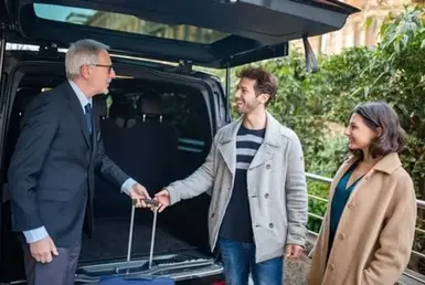 A professional driver in uniform helps a couple unload their luggage from a luxurious black car after they appear to have arrived at their destination from the airport. Representing Vancouver Airport professional chauffeur services.