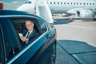 A businessman smiles while checking his smartphone in the backseat of a black luxury car driven by a chauffeur, symbolizing a professional Vancouver (VYR) transfer service.