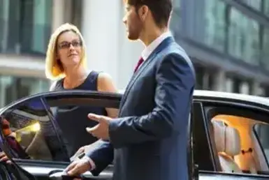 A professional chauffeur opens the door of a black luxury car to greet a businesswoman, representing Luxury Black Car Service in Houston.