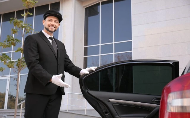 An elegantly attired man wearing a hat stands alongside a car, suggesting the presence of a chauffeur service in the vicinity.