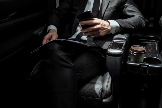Businessman on black suit sittin in a car holding smartphone on the way to meeting.