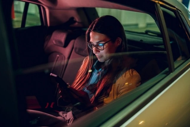 A businesswoman in glasses sits in the back seat of a car, using her smartphone to send emails or messages during a nighttime business trip.