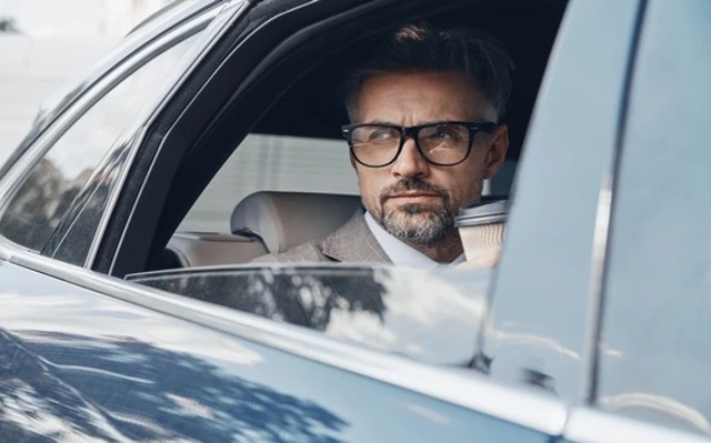A confident mature businessman, wearing glasses and a suit, is seated in the driver's seat of a car.