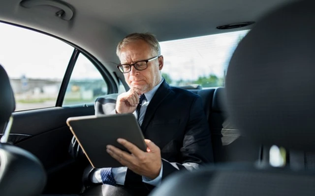 A Elderly businessman in a car using a tablet while enjoying the comfort of Private Chauffeur Service: Elevating Elderly Transportation.