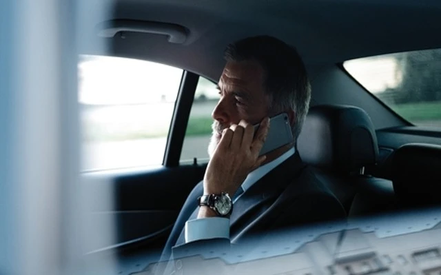 A man in a suit and tie is seated in the back seat of a car, engaged in a phone conversation. This image depicts an elderly man utilizing a cell phone while commuting for the Long Distance Elderly Transportation service.