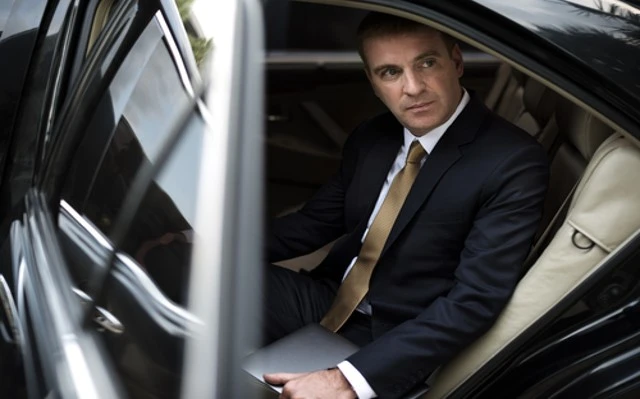 A man in a suit and tie seated in the back seat of a car, representing a businessman utilizing the Corporate Taxi Transport Service, a professional chauffeur service.