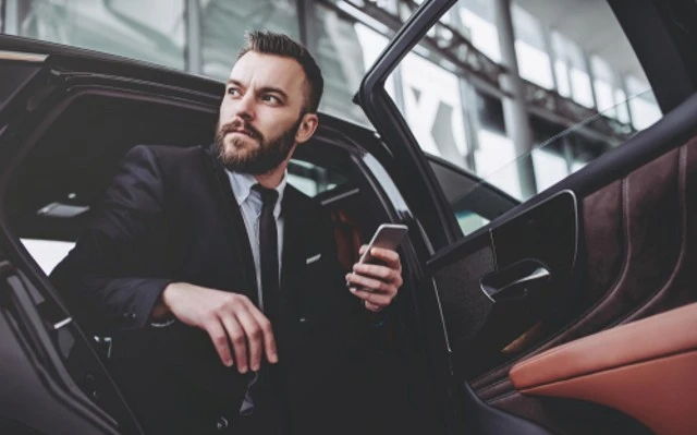 A young, handsome businessman in a suit and tie is engrossed in his smartphone while seated in the back seat of a luxury car. The serious, bearded man exudes professionalism as he utilizes roadshow services for corporate event transportation.