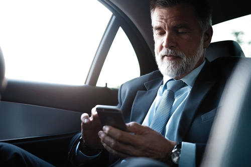 A mature business man, dressed in a suit, is seated in the back seat of a car. He is smiling and using a smartphone while enjoying the comfort of a business car. This image represents corporate event transportation services and black car service.
