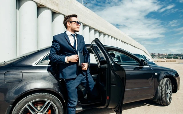 A young businessman disembarks from his black car service, wearing a suit and sunglasses. He stands confidently next to his vehicle, which is parked near a town car service. Private car service is also available nearby.