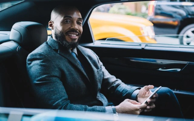 African American businessman in formal attire smiling while using cellphone in the back seat of a car during a business trip.